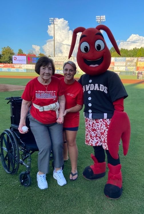 Senior woman standing up from wheelchair with young woman supporting her and Hickory Crawdads mascot Conrad the Crawdad